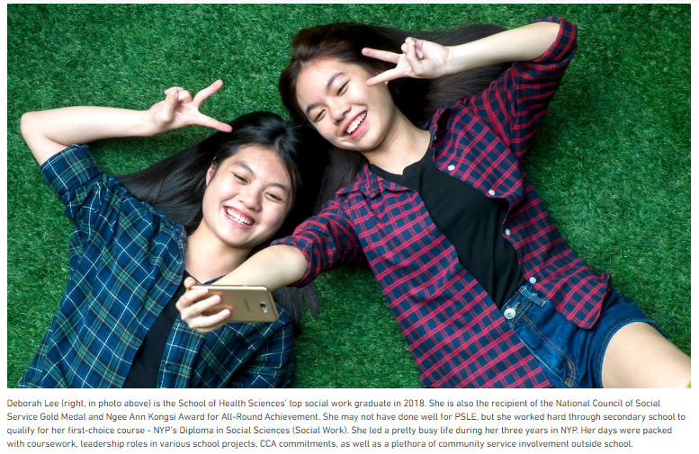 Two smiling girls lying on grass, making peace signs, taking a selfie with a gold smartphone.